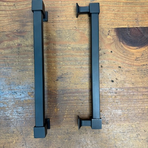 May include: Two black metal door pulls with a square design. Each pull has a rectangular bar with square ends and a small, square mounting plate. The pulls are displayed on a wooden surface.