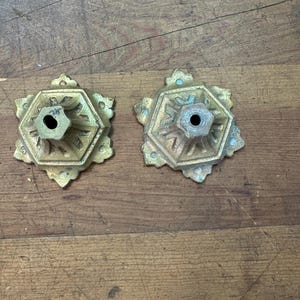 Puede incluir: Dos piezas de herrajes decorativos antiguos de latón. Cada pieza tiene una base hexagonal con detalles ornamentados y un agujero central. El latón tiene un aspecto deslustrado y envejecido, con toques de oxidación verde. Las piezas están sobre una superficie de madera.