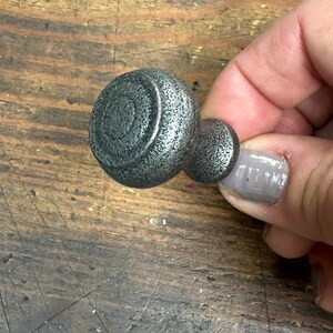 May include: A pair of dark gray metal cabinet knobs. The larger knob has a textured surface with a spiral design in the center. The smaller knob is attached to the larger one. The knobs are held by a hand.