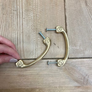 May include: Two gold-colored cabinet pulls with a floral design on each end. The pulls are curved and include screws for installation. The pulls are shown on a light-colored wooden surface.
