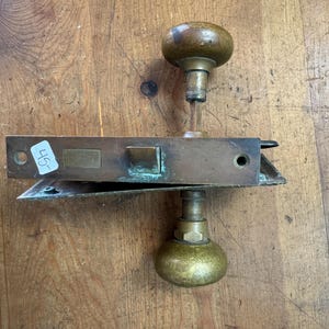 May include: Antique brass door hardware set, featuring two rounded doorknobs and a rectangular lock mechanism. The lock has a small rectangular plate and a sticker with the number "45-". The hardware shows signs of age.