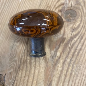 May include: A round, mushroom-shaped cabinet knob featuring a glossy, marbled finish with swirling patterns of dark brown and golden amber tones. The knob is attached to a short, cylindrical, dark metallic base with a weathered, textured surface.