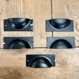 May include: Five black metal cabinet pulls with a rounded, recessed design. The pulls are rectangular in shape and have two small holes for mounting.