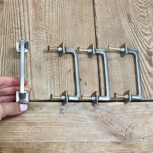 May include: A set of silver-toned cabinet pulls with a square base and a rectangular handle. The pulls are displayed on a wooden surface. One pull is held by a hand, while the others are arranged in a row, each with a gold-colored screw.