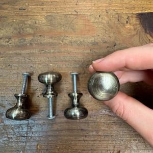 May include: A collection of antique brass cabinet knobs and pulls. The knobs have a rounded, mushroom-like shape, while the pulls have a more elongated design. The hardware is displayed on a weathered wooden surface.
