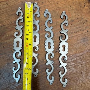 May include: Four silver-colored, ornate metal escutcheons with keyhole cutouts. Each piece is approximately 7 inches long, featuring a scrolling design. The escutcheons are displayed on a wooden surface, with a measuring tape alongside.