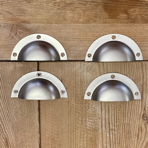 May include: Four brushed nickel half-moon cabinet pulls. Each pull has a rounded, cup-like design with three screw holes on each end. The pulls are arranged on a light brown wooden surface.