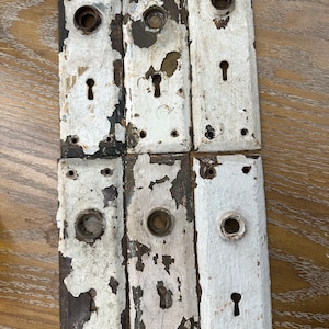 May include: Six antique door plates with keyholes and knob holes. The plates are rectangular with a distressed white paint finish over dark metal. The paint is chipped and peeling, revealing the metal underneath. The plates are arranged in two rows.