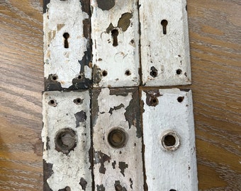 May include: Six antique door plates with keyholes and knob holes. The plates are rectangular with a distressed white paint finish over dark metal. The paint is chipped and peeling, revealing the metal underneath. The plates are arranged in two rows.