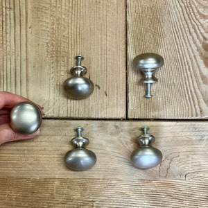 May include: Five silver cabinet knobs with a round, slightly domed shape. The knobs have a brushed finish and are mounted on a wooden surface.