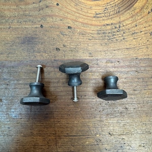 May include: Three black metal cabinet knobs with varying geometric designs. Each knob has a threaded screw for attachment. The knobs are displayed on a wooden surface with visible grain.