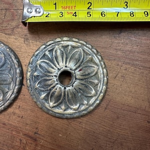 Puede incluir: Dos medallones florales antiguos de latón con un agujero central, junto a una cinta métrica. Los medallones tienen aproximadamente 5 cm de diámetro. La cinta métrica muestra medidas en pulgadas y pies.