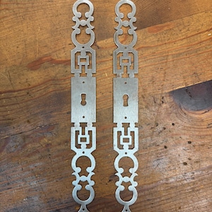 May include: Two ornate, silver-colored metal door plates with keyholes and decorative cutouts. The plates have a vertical, symmetrical design with repeating geometric and floral-inspired shapes. They are displayed on a rustic, wooden surface.