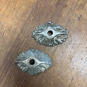 Puede incluir: Dos rosetas decorativas antiguas de metal plateado con diseños ornamentados de hojas y flores. Cada una tiene un agujero central. Las rosetas tienen forma ovalada y están sobre una superficie de madera.