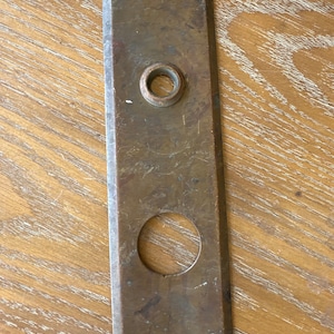 May include: A rectangular, antique brass door plate with a circular knob opening and a larger circular opening below. The plate has four screw holes and shows signs of age and wear, with a patinaed finish.