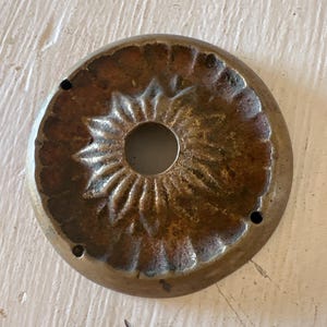 May include: A circular, antique brass ceiling medallion with a central hole and a detailed floral pattern. The medallion has a weathered, aged appearance with visible wear and tear, and small holes around the perimeter.