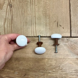 May include: Three white ceramic cabinet knobs with brown wooden bases and silver screws. One knob is round, and two are oval-shaped. The knobs are displayed on a light brown wooden surface.