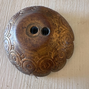 Puede incluir: Un botón vintage de color bronce con un diseño floral ornamentado. El botón es redondo con dos agujeros en el centro. Los bordes tienen un diseño festoneado. El botón tiene un acabado envejecido, lo que le da un aspecto rústico.