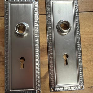 May include: Two rectangular silver-toned metal door plates with a brushed finish. Each plate features a central circular knob opening, a lower keyhole cutout, and a decorative border with a repeating scalloped pattern along the outer edges.
