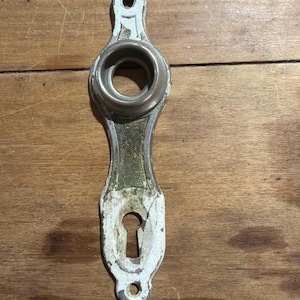 May include: A vertical metal door escutcheon plate featuring a circular knob housing at the top and a keyhole opening at the bottom. The hardware has a weathered white painted finish with visible metallic texture and two screw holes for mounting.