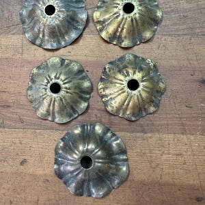 Puede incluir: Cinco cubiertas de rosetón de techo antiguas de latón con un borde con volantes y un agujero central. Las cubiertas tienen un aspecto deslustrado y envejecido, con variaciones de color de dorado a marrón oscuro. Están dispuestas sobre una superficie de madera.
