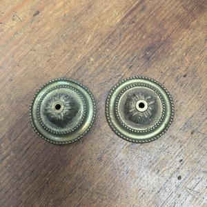 Puede incluir: Dos piezas de herrajes decorativos antiguos de latón. Cada pieza circular presenta un orificio central, un diseño floral detallado y un borde exterior con cuentas. El latón tiene una pátina cálida y envejecida.