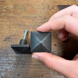 May include: Two dark gray, square cabinet knobs with a pyramid design. One knob has a screw attached. The knobs are made of metal and are displayed on a wooden surface.