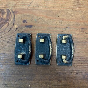 May include: Three antique-style metal drawer pulls. Each pull has a rectangular, textured black base with a gold-colored handle and two gold-colored rivets. The pulls are arranged on a wooden surface.