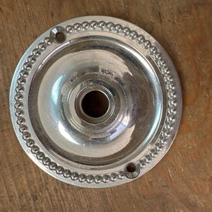 May include: A silver-colored, circular ceiling plate with a central hole and two smaller holes. The plate has a beaded edge and a reflective surface. The item is likely for electrical or lighting fixtures.