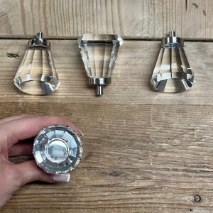 May include: Four clear crystal cabinet knobs with silver metal bases. The knobs are shaped like a diamond and have a faceted design.