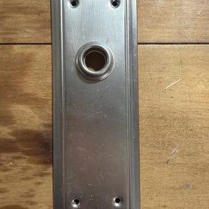 May include: A rectangular brushed nickel door plate featuring a central circular opening for a knob or spindle. The plate has a beveled edge and four small mounting holes, two positioned at the top and two at the bottom for secure installation.