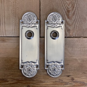 Könnte beinhalten: Zwei antike Türplatten mit verzierten floralen Mustern und einem Schlüsselloch in der Mitte. Die Platten sind aus silberfarbenem Metall gefertigt und haben eine gebürstete Oberfläche.