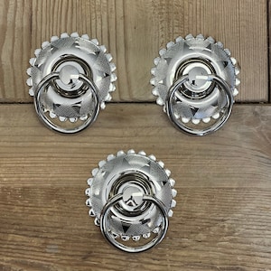 May include: Three shiny silver cabinet pulls with a decorative, patterned design. Each pull features a circular base with a ring handle. The bases have a textured pattern and a scalloped edge. The pulls are set against a wooden background.
