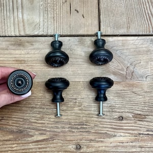 May include: A collection of black cabinet knobs with decorative detailing. One knob is held by a hand, showcasing a bronze-colored rope-like edge. The knobs are set against a rustic wooden background, highlighting their design.