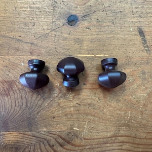 May include: Three dark brown wooden cabinet knobs with a mushroom-like design are arranged on a wooden surface. The knobs have a rounded top and a cylindrical base. The wood grain of the surface is visible.