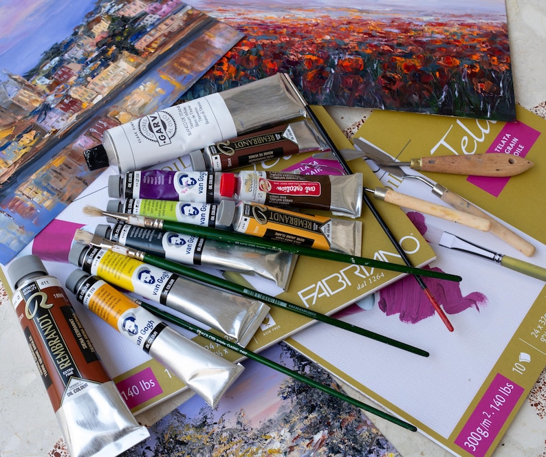 May include: A collection of oil paint tubes from brands including Rembrandt, Van Gogh, and Garv, alongside several paintbrushes with green handles, a palette knife, and a pad of Fabriano canvas paper featuring a 300 g/m2 and 140 lbs label.