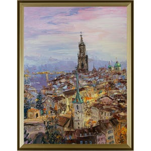 May include: An oil painting in a gold frame depicting a European cityscape at dusk. The scene features a tall church spire rising above dense buildings with warm yellow lights, set against a soft pink and lavender sky with visible brushstrokes.