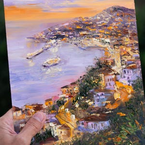 Puede incluir: Una pintura de una ciudad costera con un puerto y barcos. La ciudad est&aacute; construida en una ladera y los edificios est&aacute;n pintados en tonos de amarillo, naranja y marr&oacute;n. El cielo es de un suave p&uacute;rpura y naranja.