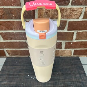 Op de afbeelding: Een cr&egrave;mekleurige waterfles met een roze handvat en de naam "Vanessa" erop gedrukt. De fles heeft een drukknopdeksel en een beige body. De handgreep is geel en roze.