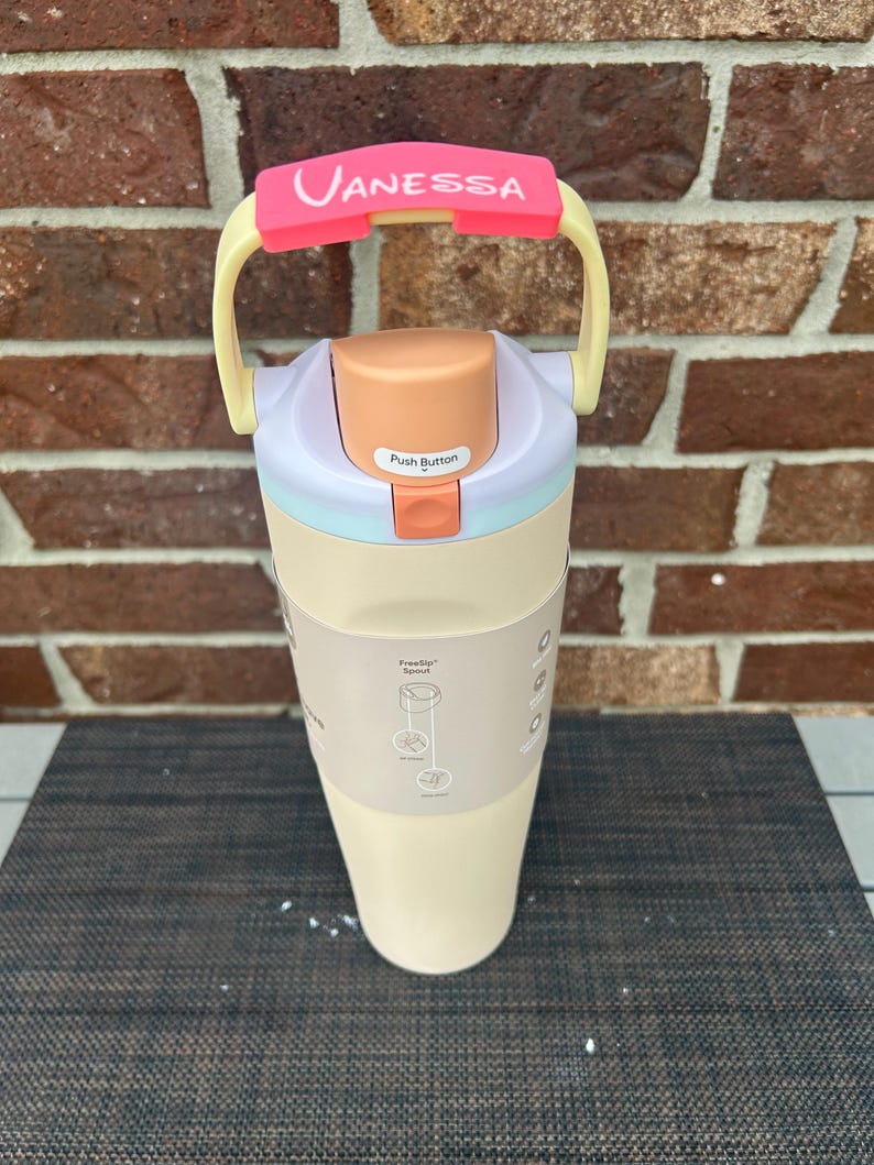 Op de afbeelding: Een cr&egrave;mekleurige waterfles met een roze handvat en de naam "Vanessa" erop gedrukt. De fles heeft een drukknopdeksel en een beige body. De handgreep is geel en roze.