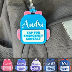 May include: A blue and pink backpack-shaped tag with the name "Audri" in white script. The tag reads "TAP FOR EMERGENCY CONTACT" in white block letters. Additional tags with names Alyssa, Vanessa, Brooke, Sawyer, and Kaj are also shown.