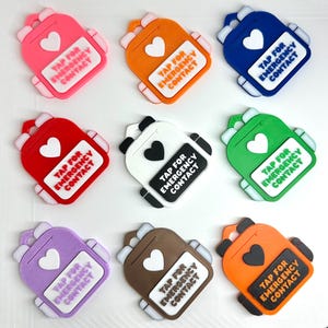May include: A collection of nine colorful backpack-shaped charms. Each charm features a heart and a rectangular label with the text "TAP FOR EMERGENCY CONTACT". The charms are in various colors, including pink, orange, blue, red, white, green, purple, brown, and orange.