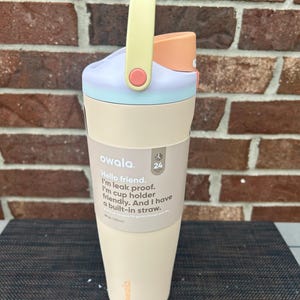Op de afbeelding: Een pastelkleurige Owala waterfles met een handvat. De fles heeft een lichtbeige basis, een lichtblauwe en paarse deksel en een perzikkleurige tuit. De handgreep is geel met een roze accent. De tekst op de fles luidt "Hello friend. I'm leak proof. I'm cup holder friendly. And I have a built-in straw."