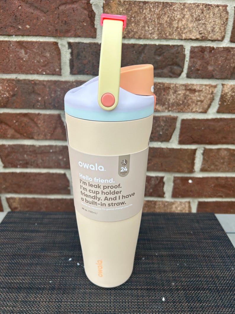 Op de afbeelding: Een pastelkleurige Owala waterfles met een handvat. De fles heeft een lichtbeige basis, een lichtblauwe en paarse deksel en een perzikkleurige tuit. De handgreep is geel met een roze accent. De tekst op de fles luidt "Hello friend. I'm leak proof. I'm cup holder friendly. And I have a built-in straw."