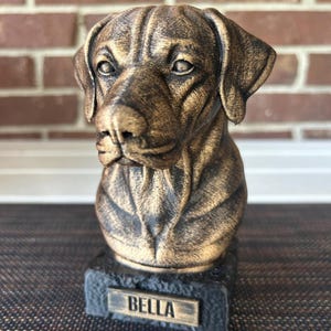 May include: A bronze-colored dog bust sculpture with a textured finish. The dog's name, "BELLA," is displayed on a small rectangular plaque at the base. The sculpture is set against a brick background.