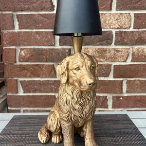 May include: A table lamp featuring a gold-colored, sculpted figure of a sitting dog. The dog has detailed fur texture and a realistic facial expression. A small, tapered gold-colored stem extends from the top of the dog's head to support a black lampshade.