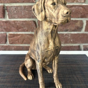 May include: A bronze-colored dog figurine in a seated position, featuring detailed fur texture and a realistic facial expression. The sculpture has a weathered, metallic finish with dark highlights that accentuate the muscular build of the animal.