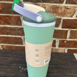 Op de afbeelding: Een mintgroene reismok met een witte en groene deksel, een lavendelkleurige handgreep en een groene sluiting. De beker heeft een witte band in het midden en een papieren hoes met tekst. BPA-vrij en gemakkelijk schoon te maken.