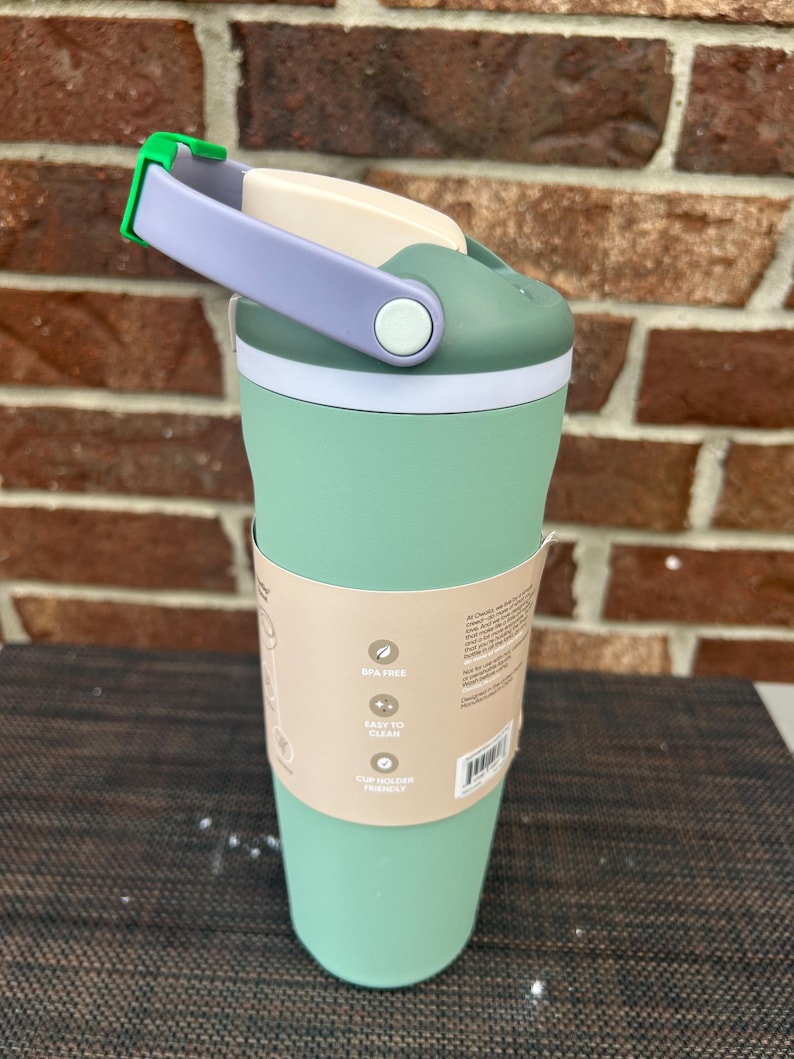 Op de afbeelding: Een mintgroene reismok met een witte en groene deksel, een lavendelkleurige handgreep en een groene sluiting. De beker heeft een witte band in het midden en een papieren hoes met tekst. BPA-vrij en gemakkelijk schoon te maken.