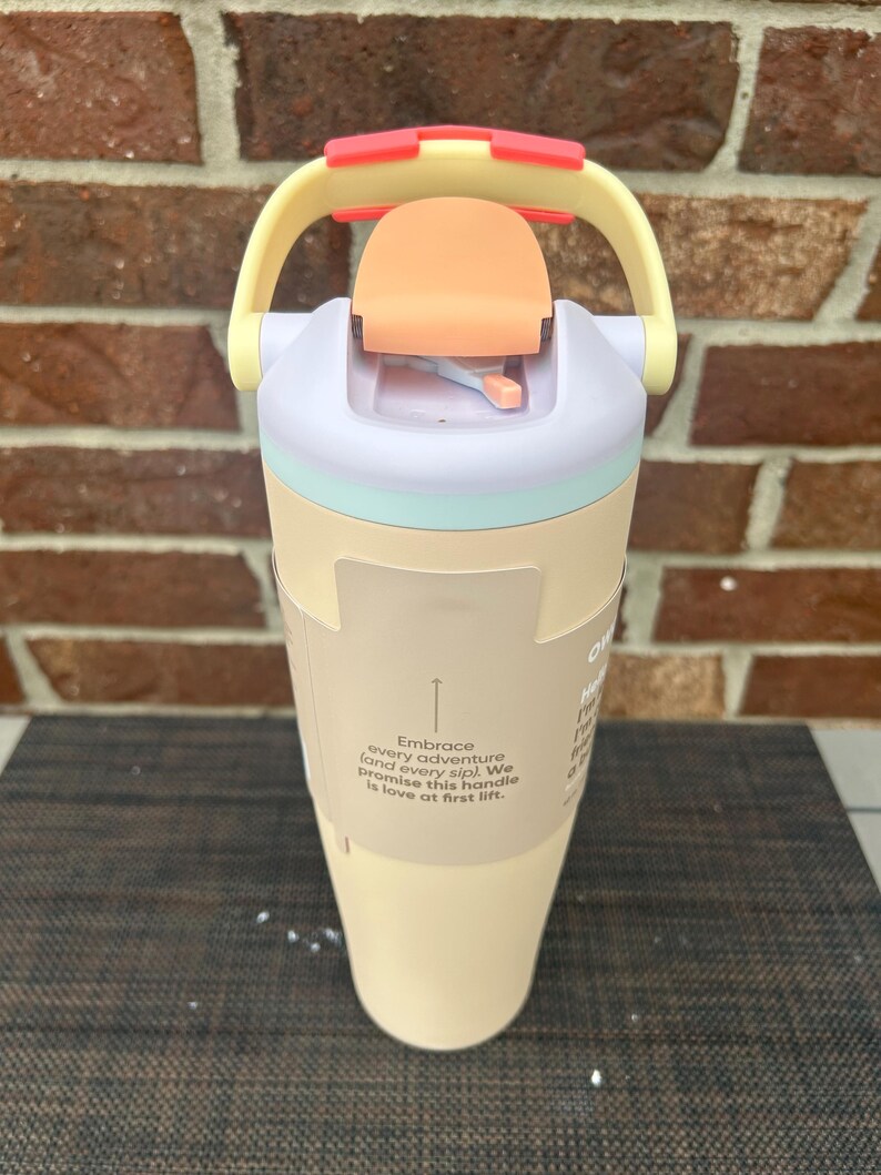 Op de afbeelding: Een pastelkleurige waterfles met een handvat en een klapdeksel. De fles is voornamelijk cr&egrave;mekleurig met een lichtblauwe band, een perzikkleurig deksel en een gele handgreep met rode accenten. De tekst op de fles luidt "Embrace every adventure (and every sip)."